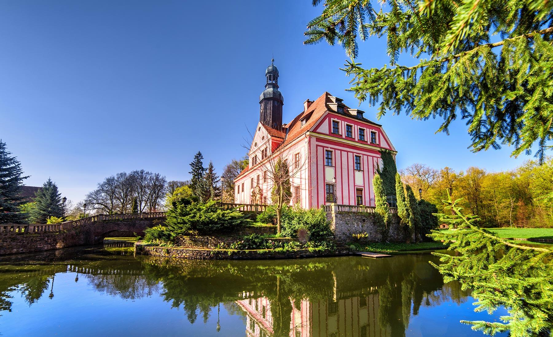 Różowa fasada Pałacu Warmątowice z wieżą odbita w wodzie fosy w słoneczny dzień